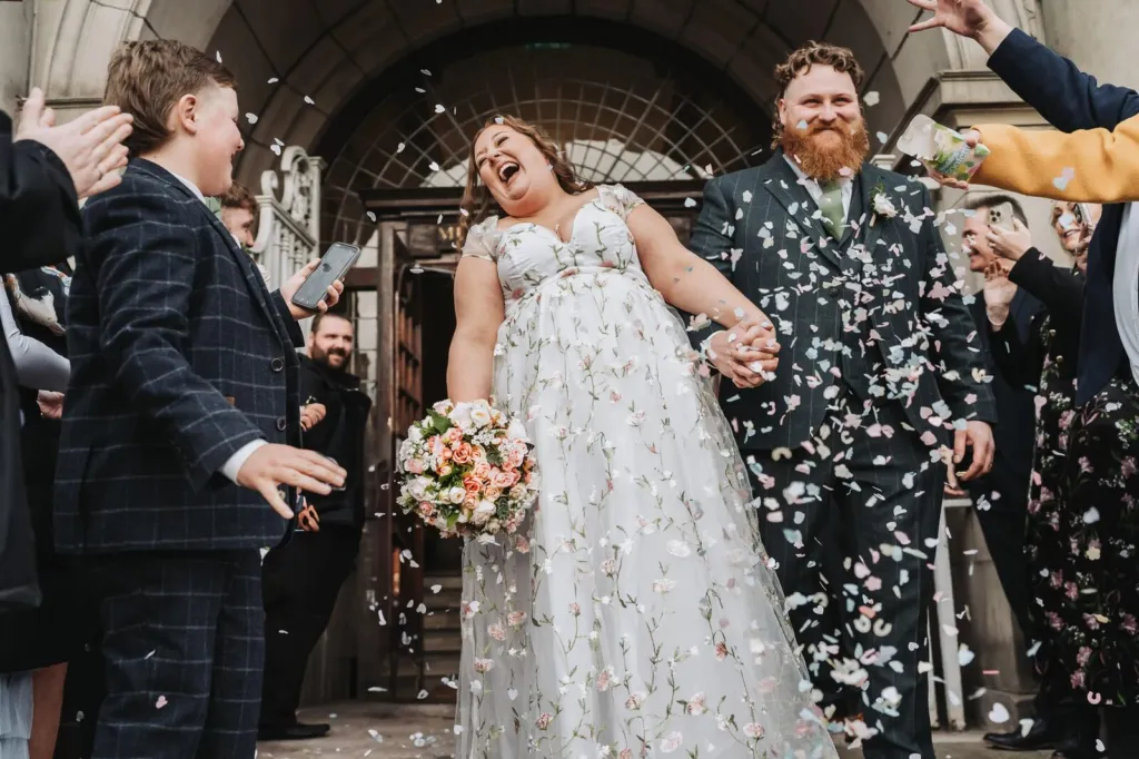 Bride in a floral gown laughing as guests throw confetti outside the ceremony doors