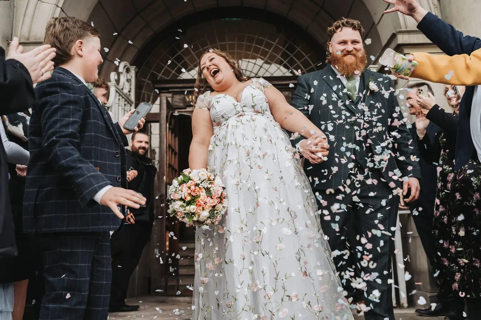 Bride in a floral gown laughing as guests throw confetti outside the ceremony doors