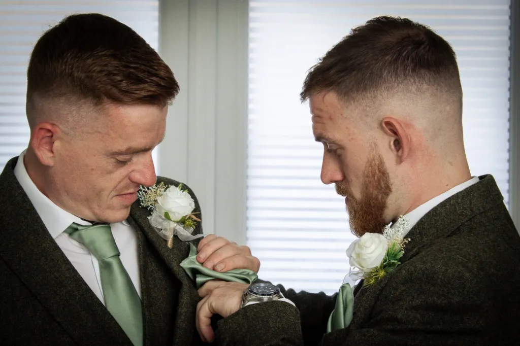 A groomsman in a tweed suit adjusting a sage green tie by a window
