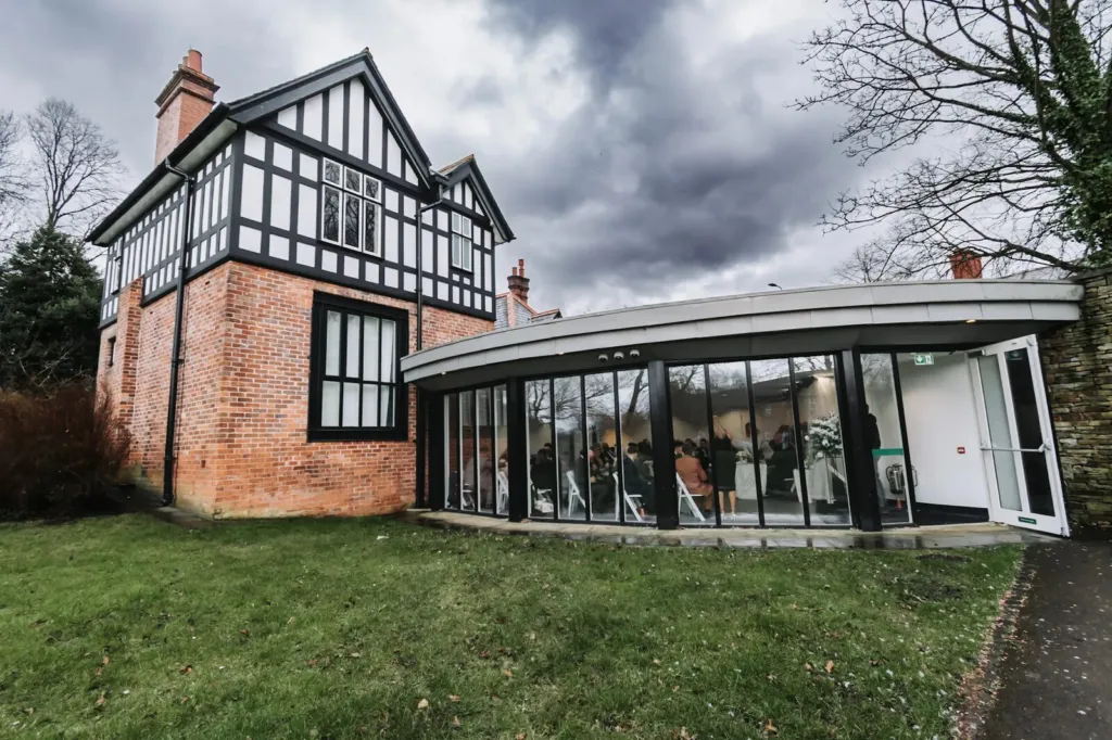A curved glass ceremony pavilion beside a Tudor house under a stormy sky