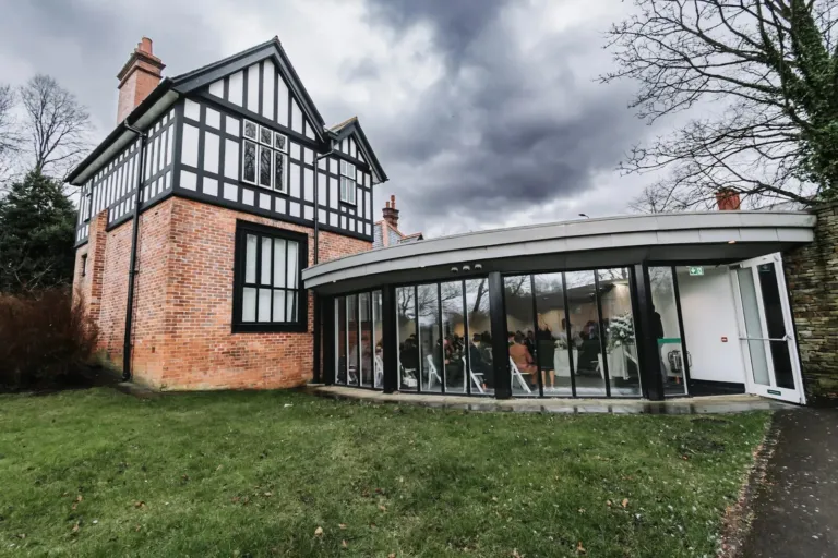 A curved glass ceremony pavilion beside a Tudor house under a stormy sky